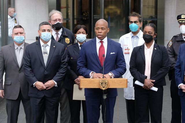 Mayor Eric Adams, wearing a red tie and navy suit and standing at a podium, holds a press conference outside New York-Presbyterian Hospital where an off-duty NYPD officer was recovering after a bullet grazed his head. Adams is flanked by several officials and doctors wearing masks.